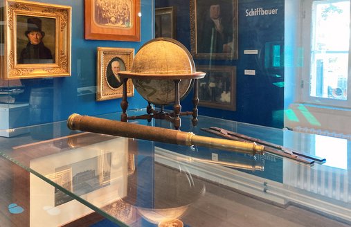 Ein Fernrohr und ein Globus liegen in einer Vitrine. An der blauen Seitenwand hängen Bilder von Menschen.