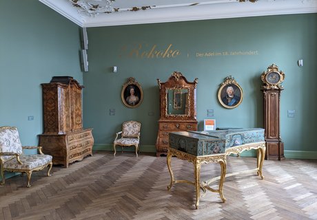 In einem Ausstellungsraum stehen Möbel aus der Zeit des Rokokos. Es gibt Holzschränke, Sessel, Bilder an der Wand und ein Cembalo. Das ist ein altes Tasteninstrument. 