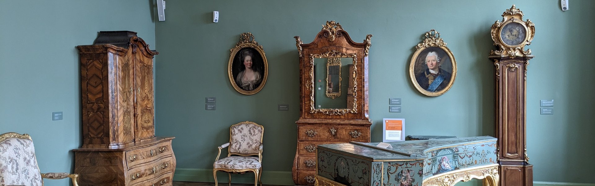  I et udstillingslokale står møbler fra rokokoperioden. Der er træskabe, stole, billeder på væggen og et cembalo. Det er et gammelt tangentinstrument. På en grøn væg står der: »Rokoko, adelen i det 18. århundrede.«