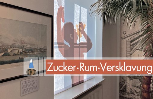 In einem Fenster der Ausstellung ZUCKER.RUM.VERSKLAVUNG. hängt eine Verdunklung, auf welcher der Freedomsfighter als Illustration abgebildet ist. In der Hand hält der Versklavte eine Machete, in der anderen das Gehäuse der Riesenfechterschnecke. In das Gehäuse ist ein Loch gebohrt, wodurch der Versklavte pustet. Auf dem Foto ist ein farbiger Balken platziert mit dem Piktogramm und dem Namen der Ausstellung.