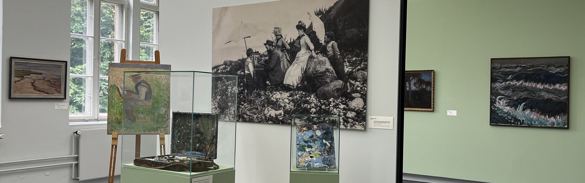 Eine bunte Fotografie. Ein Museumsraum. Im Hintergrund ist auf der linken Seite ein altes großes Fenster. Auf der rechten Seite hängen zwei Gemälde an der Wand. Im Vordergrund ist ein kleines Podest mit Wand. An der Wand hängt eine große Schwarz-weiß-Fotografie eines Mannes der im Freien an einem Bild auf einer Staffelei arbeitet. Um ihn herum stehen Frauen und Kinder in Kleidern, die man um 1900 getragen hat. Auf dem Podest ist eine Staffelei. Darauf das Bild eines Mannes beim Malen. Daneben ist eine Vitrine mit einem alten Malkoffer. Ganz rechts ist eine kleinere Vitrine mit einer alten Malerpalette.  