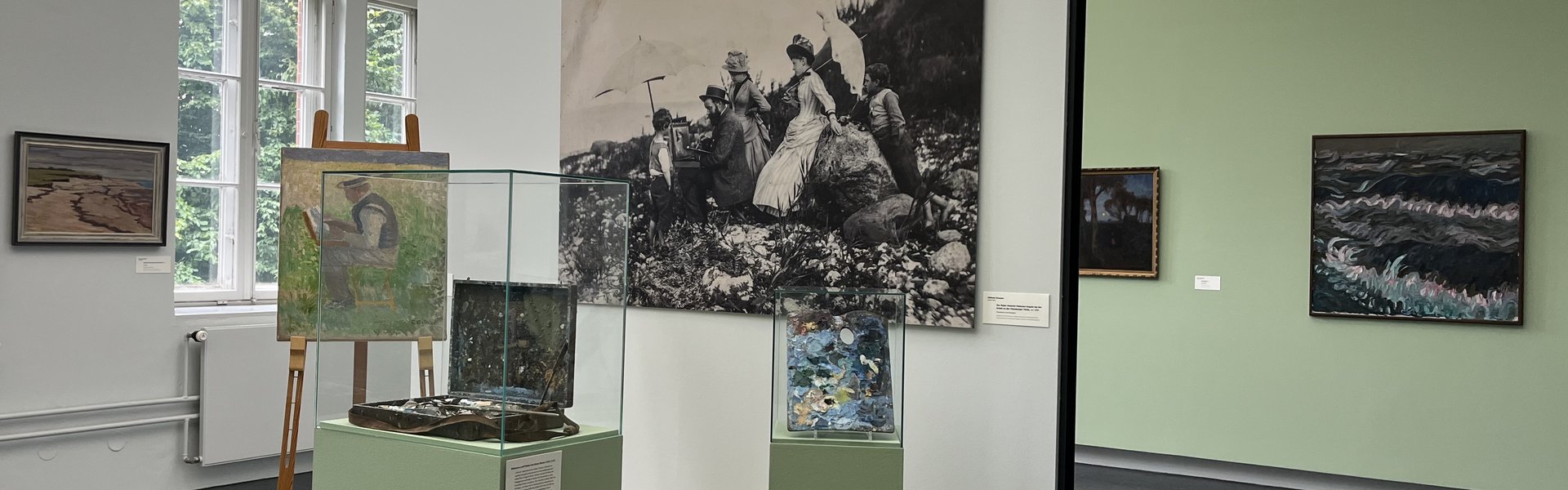 Eine bunte Fotografie. Ein Museumsraum. Im Hintergrund ist auf der linken Seite ein altes großes Fenster. Auf der rechten Seite hängen zwei Gemälde an der Wand. Im Vordergrund ist ein kleines Podest mit Wand. An der Wand hängt eine große Schwarz-weiß-Fotografie eines Mannes der im Freien an einem Bild auf einer Staffelei arbeitet. Um ihn herum stehen Frauen und Kinder in Kleidern, die man um 1900 getragen hat. Auf dem Podest ist eine Staffelei. Darauf das Bild eines Mannes beim Malen. Daneben ist eine Vitrine mit einem alten Malkoffer. Ganz rechts ist eine kleinere Vitrine mit einer alten Malerpalette.  