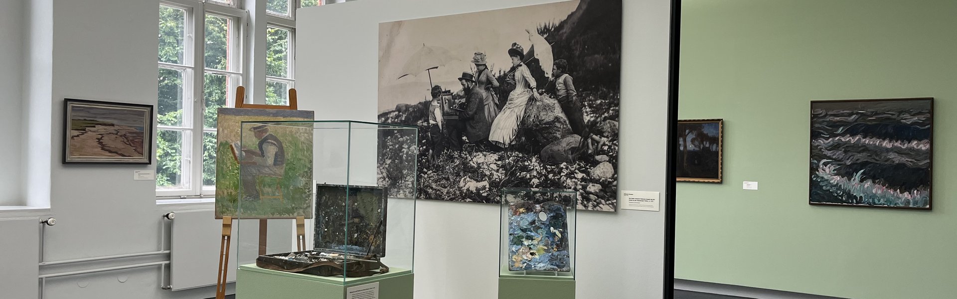 Das Bild ist eine bunte Fotografie im Museum. Es zeigt einen Museumsraum. Hinten auf der linken Seite gibt es ein großes, altes Fenster. Auf der rechten Seite hängen zwei Gemälde an der Wand. Vorne steht ein kleines Podest. Es gibt auch eine Wand. An der Wand hängt eine große Fotografie. Auf der Fotografie sieht man einen Mann, der draußen ein Bild malt. Er arbeitet an einer Staffelei. Um ihn herum stehen Frauen und Kinder. Sie tragen Kleider, die man um 1900 getragen hat. Auf dem Podest steht eine Staffelei. Auf der Staffelei ist das Bild von dem Mann, der malt. Daneben gibt es eine Vitrine. In der Vitrine liegt ein alter Malkoffer. Ganz rechts ist eine kleinere Vitrine. In dieser Vitrine liegt eine alte Malerpalette.