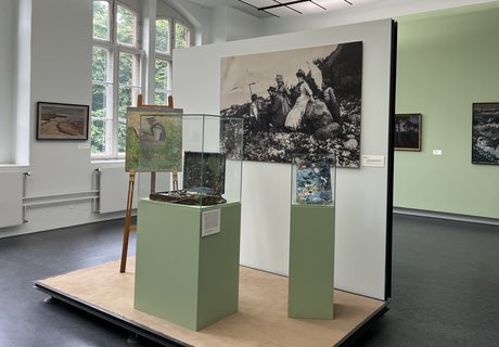 Das Bild ist eine bunte Fotografie im Museum. Es zeigt einen Museumsraum. Hinten auf der linken Seite gibt es ein großes, altes Fenster. Auf der rechten Seite hängen zwei Gemälde an der Wand. Vorne steht ein kleines Podest. Es gibt auch eine Wand. An der Wand hängt eine große Fotografie. Auf der Fotografie sieht man einen Mann, der draußen ein Bild malt. Er arbeitet an einer Staffelei. Um ihn herum stehen Frauen und Kinder. Sie tragen Kleider, die man um 1900 getragen hat. Auf dem Podest steht eine Staffelei. Auf der Staffelei ist das Bild von dem Mann, der malt. Daneben gibt es eine Vitrine. In der Vitrine liegt ein alter Malkoffer. Ganz rechts ist eine kleinere Vitrine. In dieser Vitrine liegt eine alte Malerpalette.