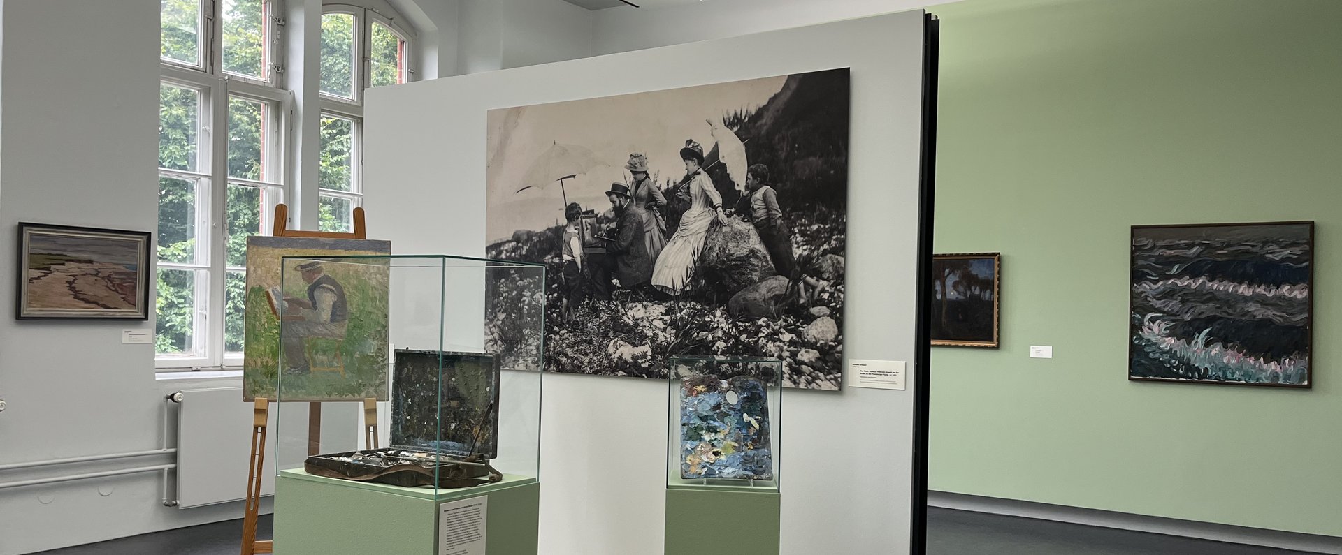 Eine bunte Fotografie. Ein Museumsraum. Im Hintergrund ist auf der linken Seite ein altes großes Fenster. Auf der rechten Seite hängen zwei Gemälde an der Wand. Im Vordergrund ist ein kleines Podest mit Wand. An der Wand hängt eine große Schwarz-weiß-Fotografie eines Mannes der im Freien an einem Bild auf einer Staffelei arbeitet. Um ihn herum stehen Frauen und Kinder in Kleidern, die man um 1900 getragen hat. Auf dem Podest ist eine Staffelei. Darauf das Bild eines Mannes beim Malen. Daneben ist eine Vitrine mit einem alten Malkoffer. Ganz rechts ist eine kleinere Vitrine mit einer alten Malerpalette.  