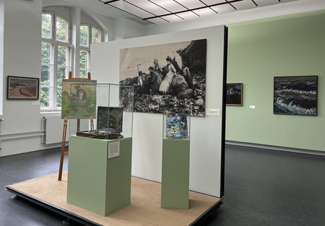 Eine bunte Fotografie. Ein Museumsraum. Im Hintergrund ist auf der linken Seite ein altes großes Fenster. Auf der rechten Seite hängen zwei Gemälde an der Wand. Im Vordergrund ist ein kleines Podest mit Wand. An der Wand hängt eine große Schwarz-weiß-Fotografie eines Mannes der im Freien an einem Bild auf einer Staffelei arbeitet. Um ihn herum stehen Frauen und Kinder in Kleidern, die man um 1900 getragen hat. Auf dem Podest ist eine Staffelei. Darauf das Bild eines Mannes beim Malen. Daneben ist eine Vitrine mit einem alten Malkoffer. Ganz rechts ist eine kleinere Vitrine mit einer alten Malerpalette.  