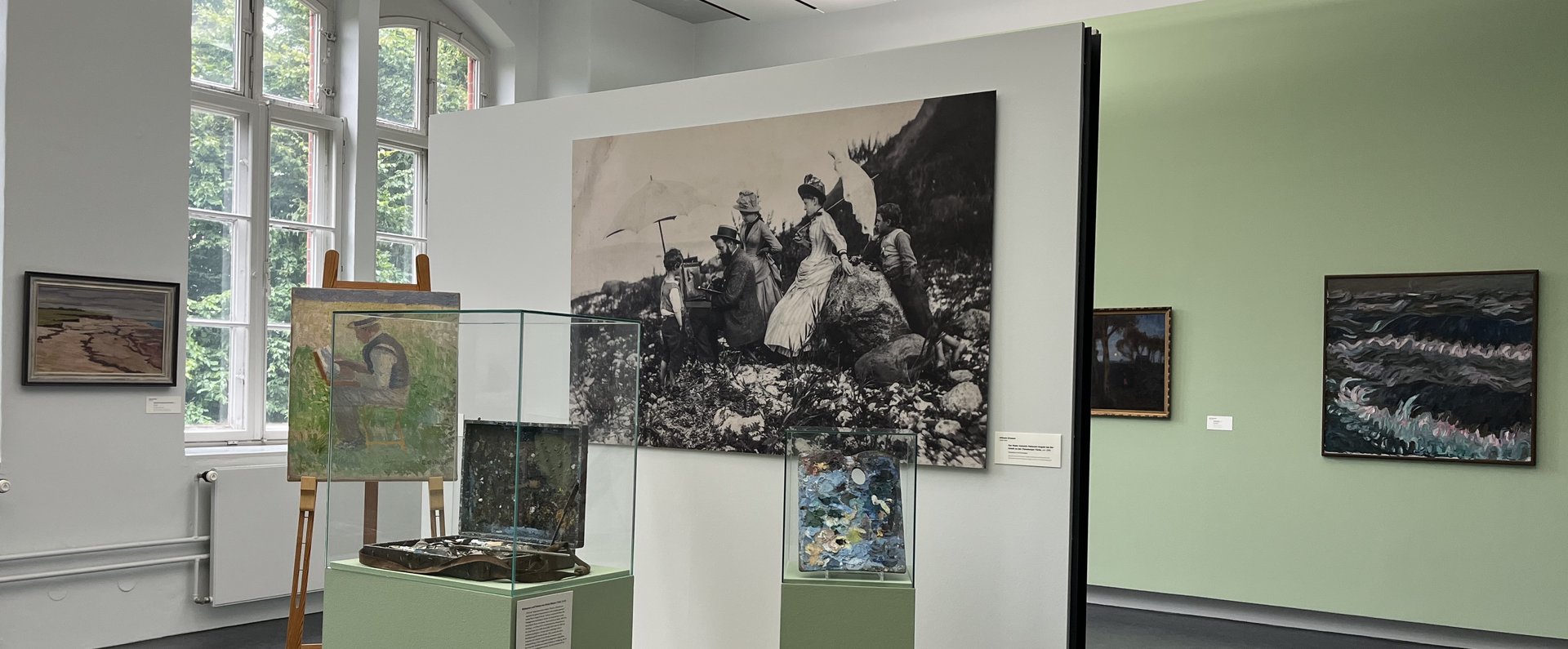 Eine bunte Fotografie. Ein Museumsraum. Im Hintergrund ist auf der linken Seite ein altes großes Fenster. Auf der rechten Seite hängen zwei Gemälde an der Wand. Im Vordergrund ist ein kleines Podest mit Wand. An der Wand hängt eine große Schwarz-weiß-Fotografie eines Mannes der im Freien an einem Bild auf einer Staffelei arbeitet. Um ihn herum stehen Frauen und Kinder in Kleidern, die man um 1900 getragen hat. Auf dem Podest ist eine Staffelei. Darauf das Bild eines Mannes beim Malen. Daneben ist eine Vitrine mit einem alten Malkoffer. Ganz rechts ist eine kleinere Vitrine mit einer alten Malerpalette.  