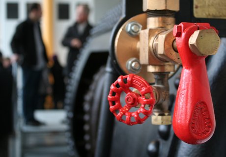 I forgrunden ses et skibs dampkedel. I baggrunden står to personer og taler sammen.