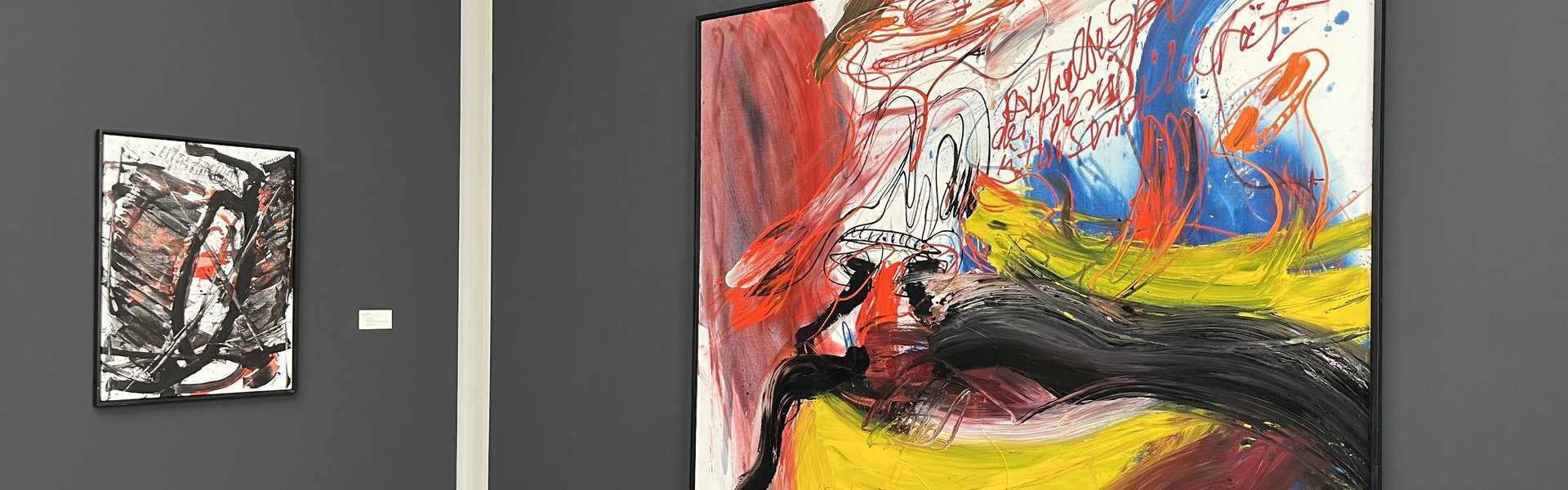 Auf der Fotografie sind zwei Gemälde vor einer grauen Wand zu sehen. Beide Bilder sind ungegenständlich. Das rechte zeigt auffällige rote, leuchtend gelbe und schwarze Farbverläufe. Das linke kräftige schwarze Linien und einige rote Farbfelder.