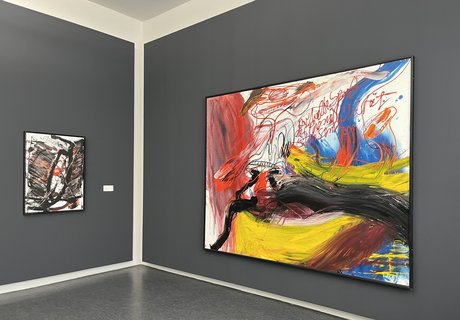 Auf der Fotografie sind zwei Gemälde vor einer grauen Wand zu sehen. Beide Bilder sind ungegenständlich. Das rechte zeigt auffällige rote, leuchtend gelbe und schwarze Farbverläufe. Das linke kräftige schwarze Linien und einige rote Farbfelder.