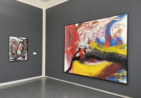 Auf der Fotografie sind zwei Gemälde vor einer grauen Wand zu sehen. Beide Bilder sind ungegenständlich. Das rechte zeigt auffällige rote, leuchtend gelbe und schwarze Farbverläufe. Das linke kräftige schwarze Linien und einige rote Farbfelder.