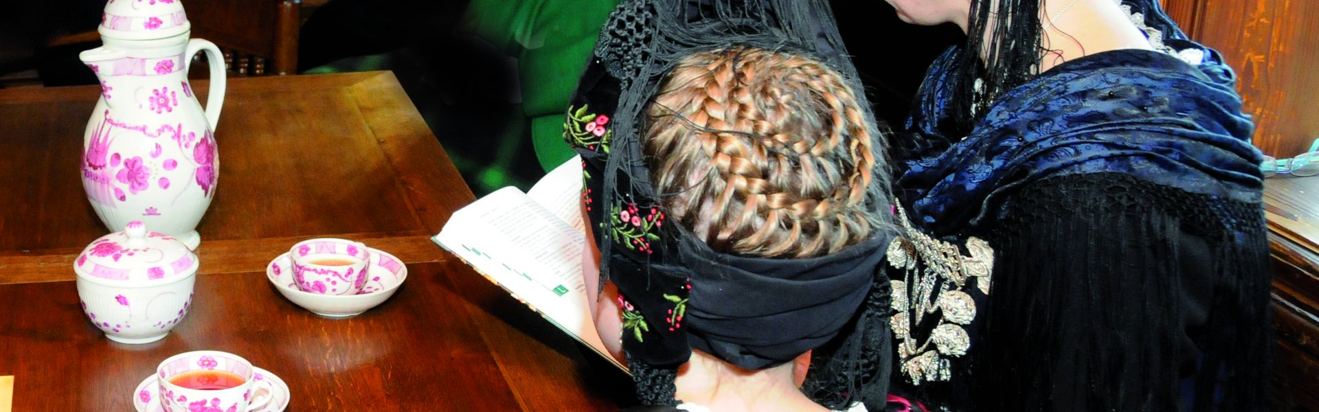 Es sind zwei Frauen in friesischer Tracht zu sehen, die Teetassen vor sich stehen haben.