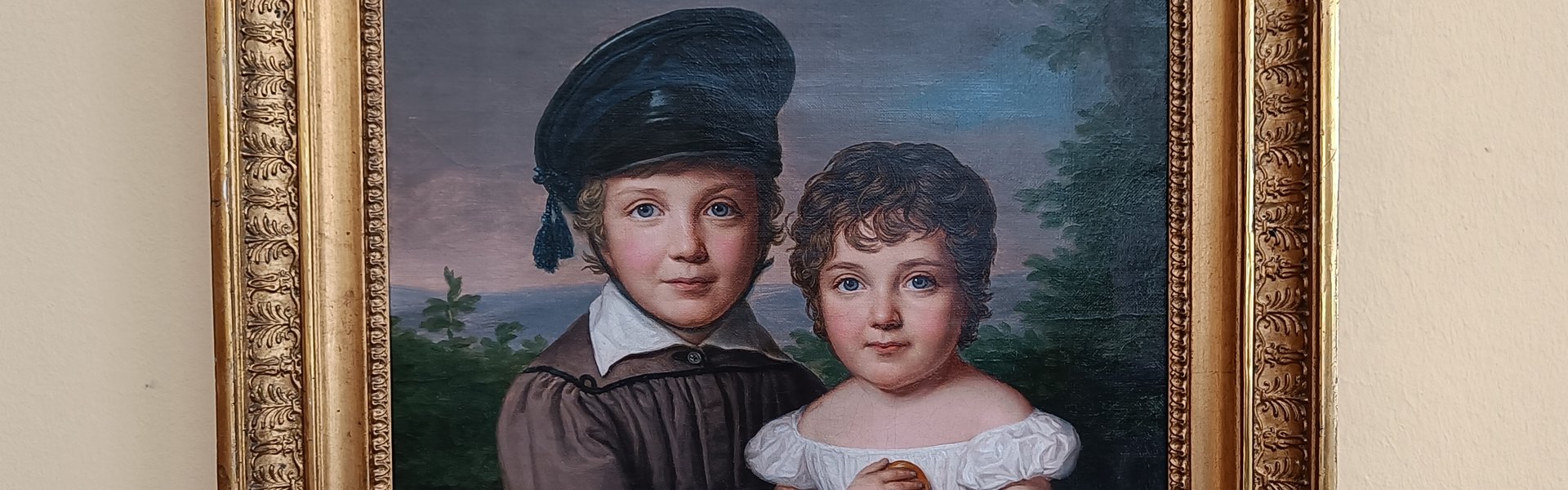 Eine bunte Fotografie. Darauf zu sehen ist eine helle Wand. Daran hängt ein Gemälde mit einem verzierten Messingrahmen. Dargestellt sind zwei Kinder. Ein Junge mit einer Mütze und in einem hellbraunen Gewand. Das Mädchen trägt ein weißes Kleid. Auf ihrem Schoß steht ein Korb mit Äpfeln. 