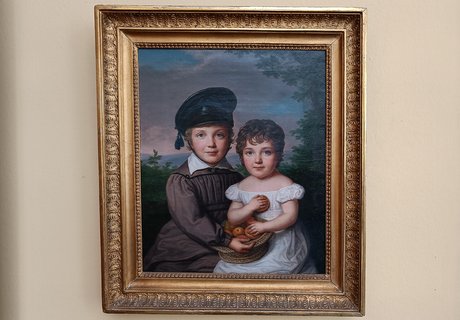 Auf der Fotografie sehen Sie eine helle Wand. An der Wand hängt ein Gemälde in einem verzierten Rahmen aus Messing. Das Gemälde zeigt zwei Kinder. Ein Junge trägt eine Mütze und ein hellbraunes Gewand. Das Mädchen trägt ein weißes Kleid. Auf ihrem Schoß steht ein Korb mit Äpfeln.