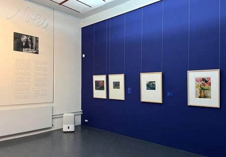 Eine bunte Fotografie. Darauf zu sehen ist die Zimmerecke eines Ausstellungsraums des Museumsberg Flensburgs. Auf der linken Seite ist ein Ausstellungstext zu sehen. Teil der Überschrift lautet „Nolde“. Über dem Text ist eine Schwarz-Weiß-Fotografie von einem Mann und einer Frau zu erkennen. Rechts davon ist die Wand in einem dunklen Blau gestrichen. An ihr hängen vier bunte Gemälde. Jedes Gemälde ist in einem hellbraunen Holzrahmen. 