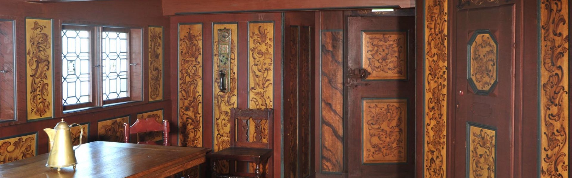 An den Wänden und der Decke des Raumes ist Holz mit Verzierungen angebracht. Ein Stuhl und ein Tisch stehen darin.