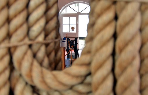 Im Vordergrund hängt ein dickes und langes Seil in Schlaufen gelegt vor der Decke herunter. Durch die Schlaufe kann man die Reepschlagmaschine sehen und die offene Holztür, die auf den Innenhof des Flensburger Schifffahrtsmuseums führt.