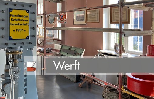 Links steht eine Maschine mit einem gelben Schild. Auf diesem steht: Flensburger Schiffsbau Gesellschaft 1957. Links daneben steht ein langes Metallregal. Von den Regelbögen hängen Bilder herunter. Hierbei handelt es sich um Schriftstücke und Impressionen von einer Werft. In den Regalen liegt unterschiedliches Werkzeug – z.B. ein Niethammer ganz hinten im Bild.