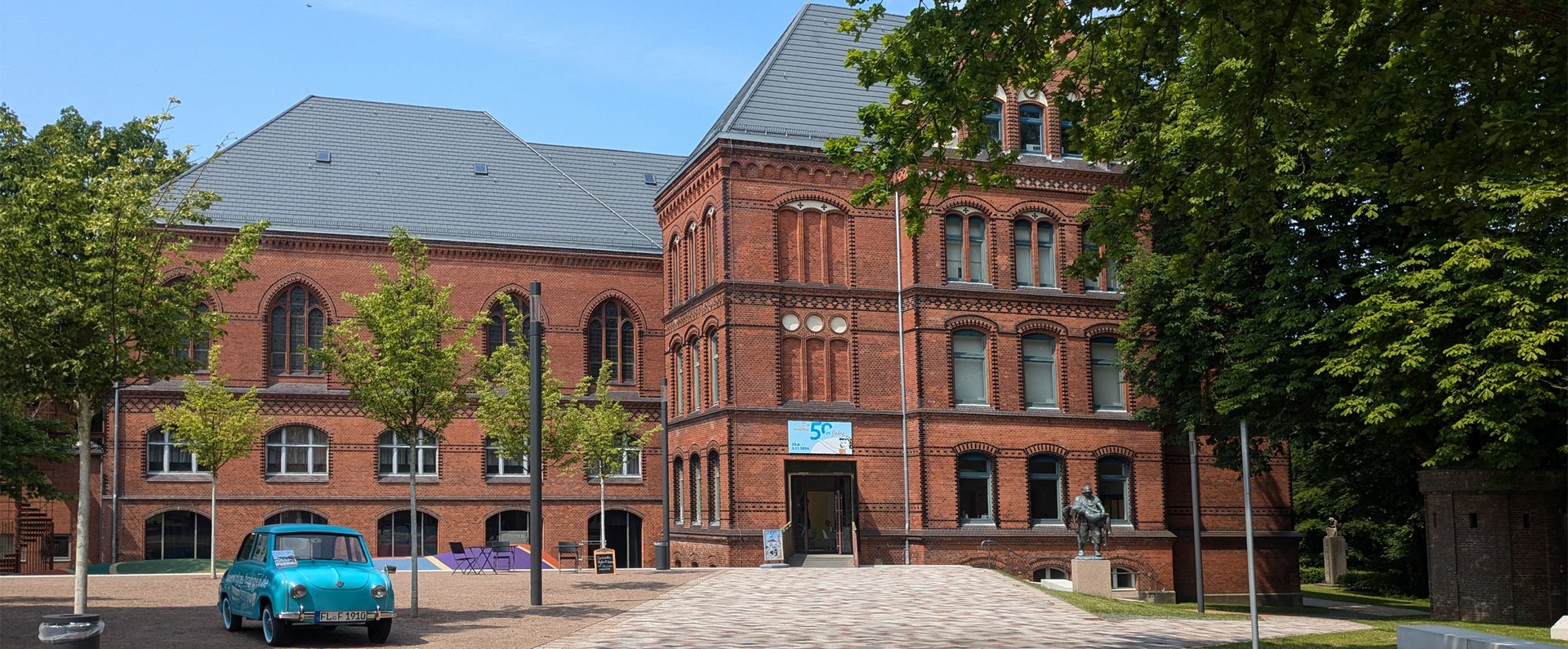 Das rote Gebäude ist das Hans-Christiansen-Haus. Davor stehen ein blaues Auto und eine Skulptur. Ein Weg führt zum Eingang. Daneben sind Bäume.