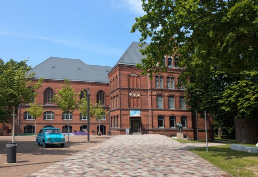Das rote Gebäude ist das Hans-Christiansen-Haus. Davor stehen ein blaues Auto und eine Skulptur. Ein Weg führt zum Eingang. Daneben sind Bäume.