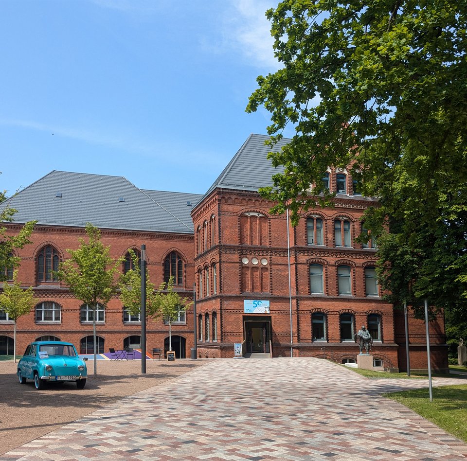 Das rote Gebäude ist das Hans-Christiansen-Haus. Davor stehen ein blaues Auto und eine Skulptur. Ein Weg führt zum Eingang. Daneben sind Bäume.