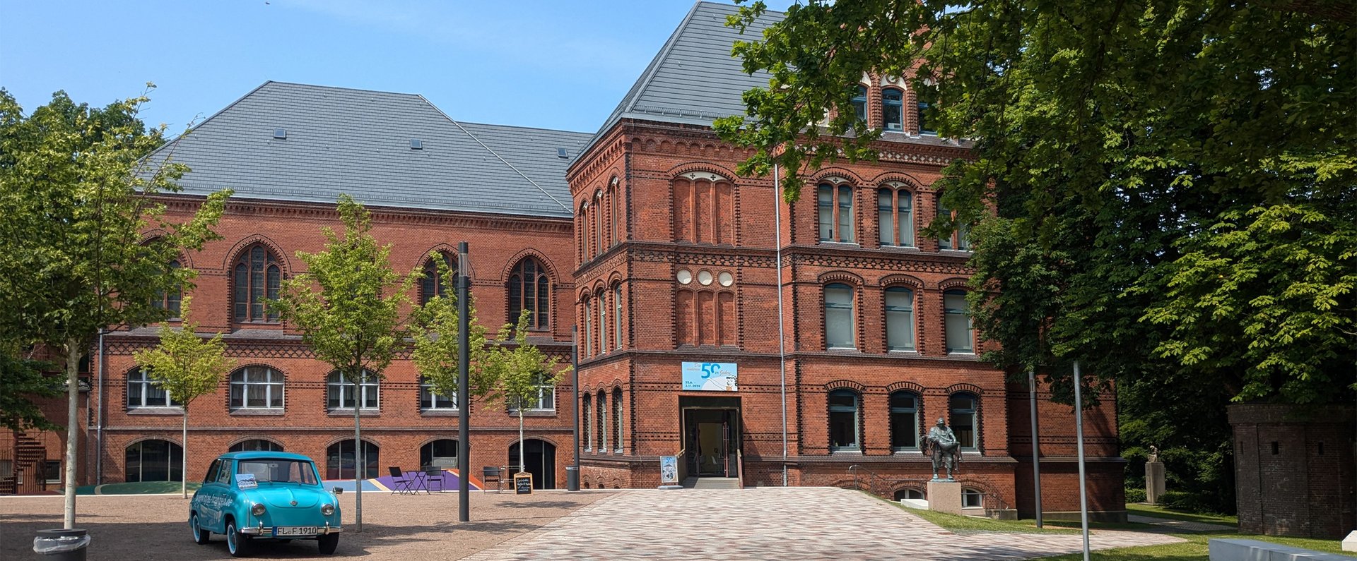 Das rote Gebäude ist das Hans-Christiansen-Haus. Davor stehen ein blaues Auto und eine Skulptur. Ein Weg führt zum Eingang. Daneben sind Bäume.