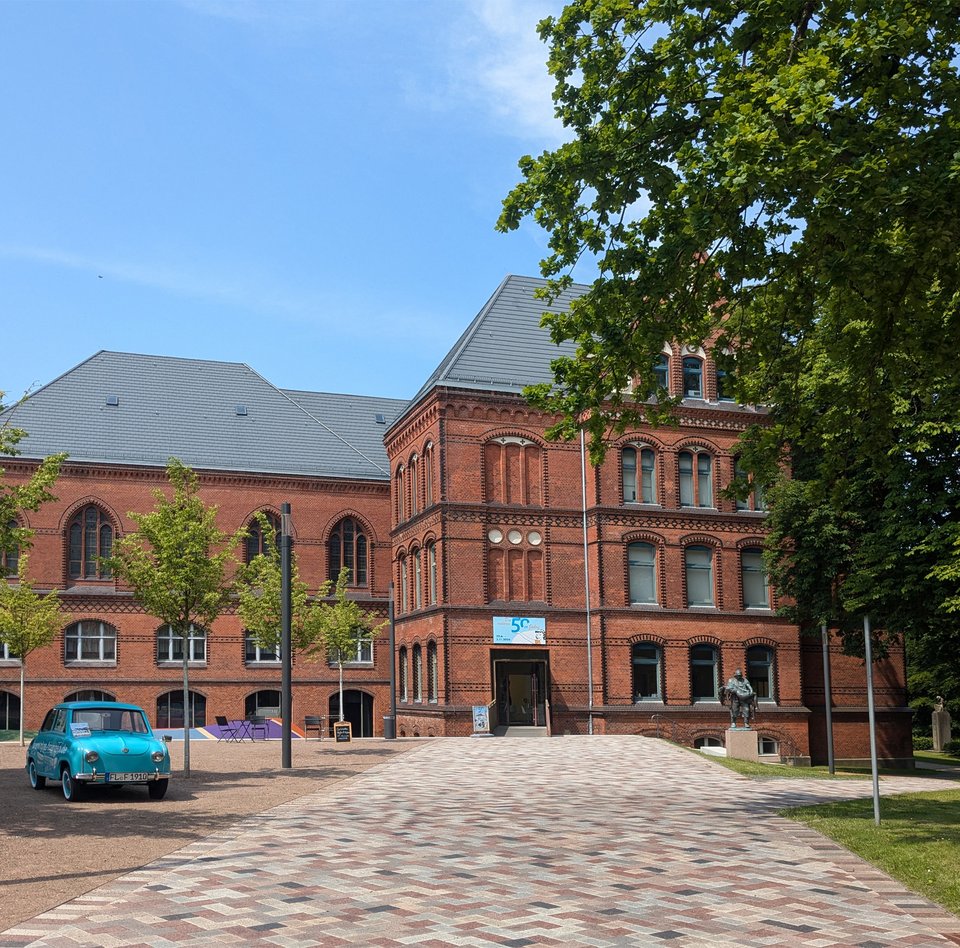 Das rote Gebäude ist das Hans-Christiansen-Haus. Davor stehen ein blaues Auto und eine Skulptur. Ein Weg führt zum Eingang. Daneben sind Bäume.