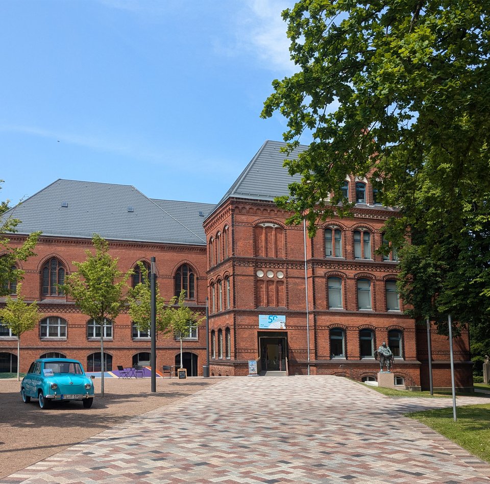 Den røde bygning er Hans Christiansen-huset. Foran den står en blå bil og en skulptur. En sti fører til indgangen. Ved siden af er der træer.