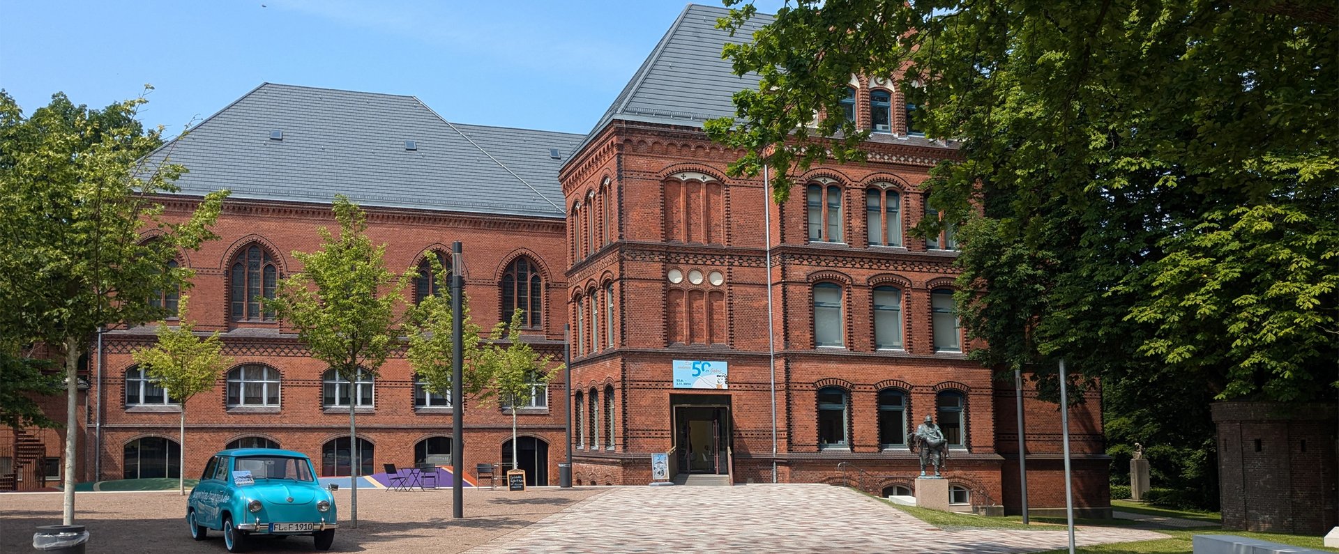  Heinrich-Sauermann-Haus er en del af Museumsberg Flensburg. Det er en bygning af røde sten. Taget er også rødt og blåt. Huset har mange dekorationer og vinduer. Omkring bygningen er der blomstrende træer og blomster. Himlen er blå.