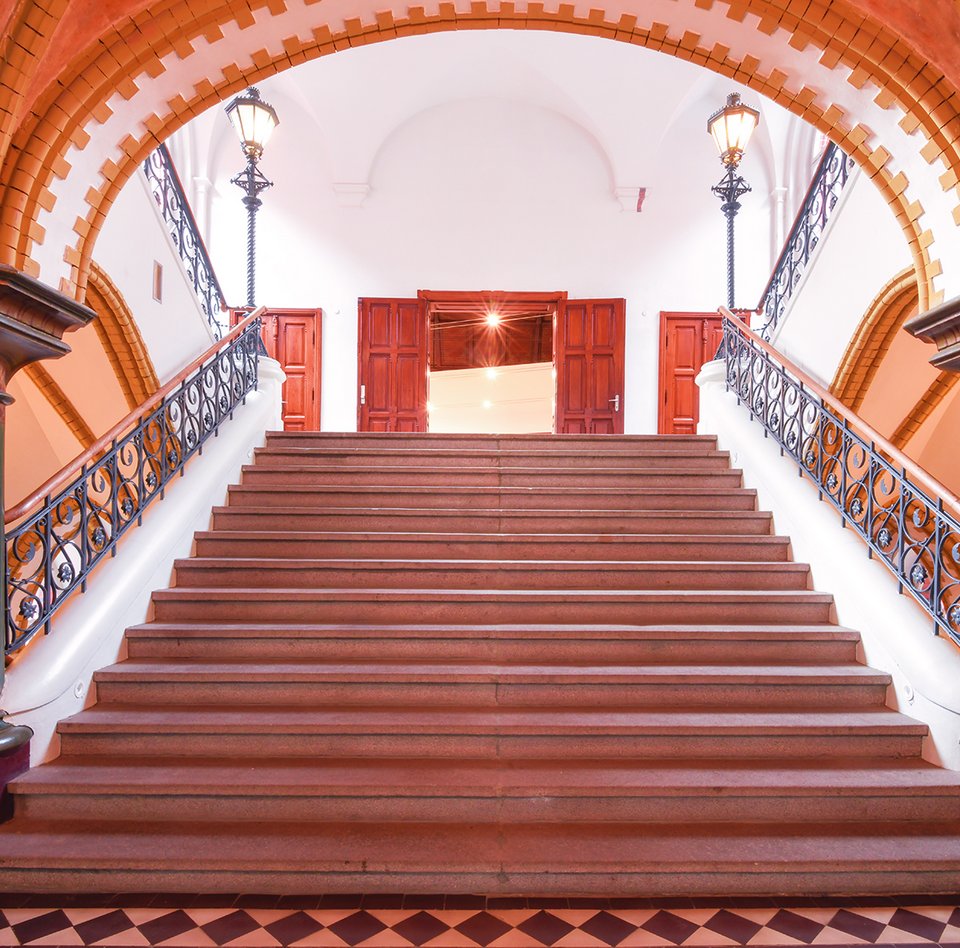 En stentrappe fører til en stor åben dør. Ved trappens begyndelse ser man to søjler og ved siden af trappeopgangen.