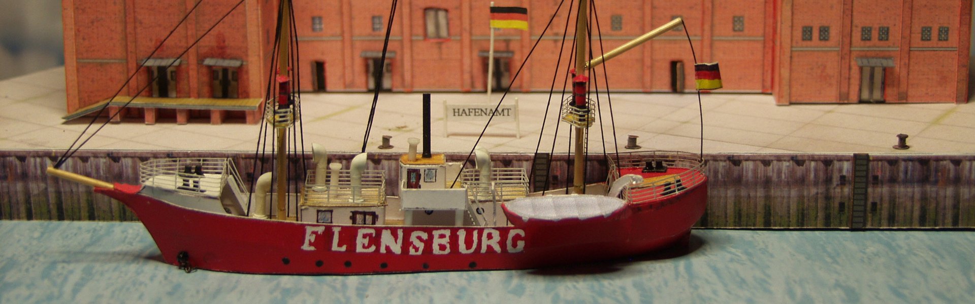 Auf dem Foto ist ein Modellschiff, das aus Karton hergestellt worden ist zu sehen. Es ist rot und trägt den Namen FLENSBURG. Es liegt an der Pier, die ebenfalls aus Pappe hergestellt worden ist. Im Hintergrund ist eine rotes, großes Gebäude zu sehen - ebenfalls aus Karton.