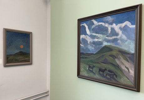 Das Foto zeigt zwei Gemälde von Käte Lassen. Sie hängen in einem Raum mit einer mintgrünen Wand. Auf dem rechten Gemälde sieht man eine hügelige Landschaft. In der Mitte des Bildes leuchtet ein orangefarbener Mond. Auf dem linken Gemälde ist auch eine hügelige Landschaft zu sehen. Über dem Horizont ist der blaue Himmel mit weißen Wolken. Vorne stehen vier Schafe.