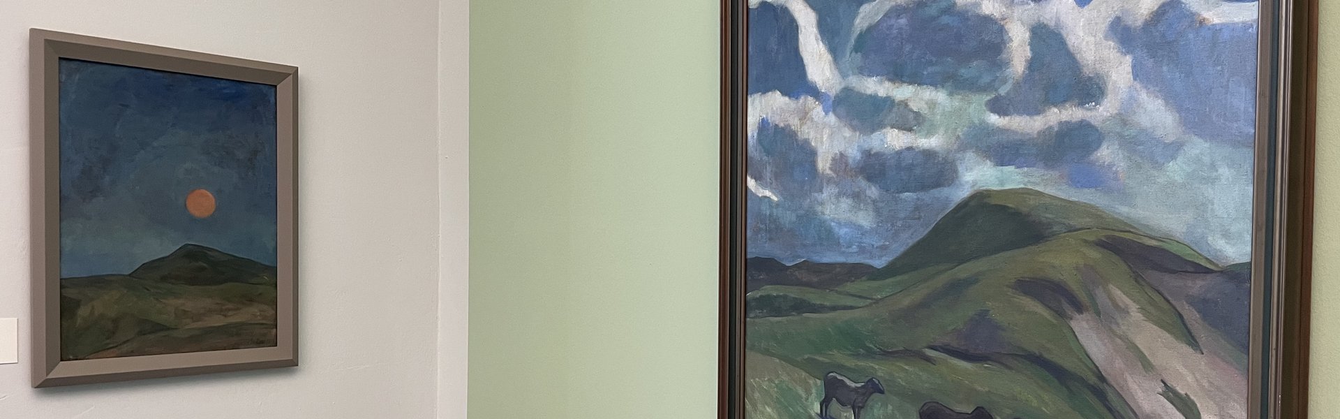 Das Foto zeigt zwei Gemälde von Käte Lassen. Sie hängen in einem Ausstellungsraum vor einer mintgrünen Wand. Auf dem rechten Gemälde ist eine hügelige Landschaft zu sehen. In der Bildmitte befindet sich ein orangefarbener, leuchtender Mond. Auf dem linken Gemälde ist ebenfalls eine hügelige Landschaft zu sehen. Über dem Horizont ist der blaue Himmel mit weißen Wolken dargestellt. In der Landschaft im Bildvordergrund befinden sich vier Schafe.  