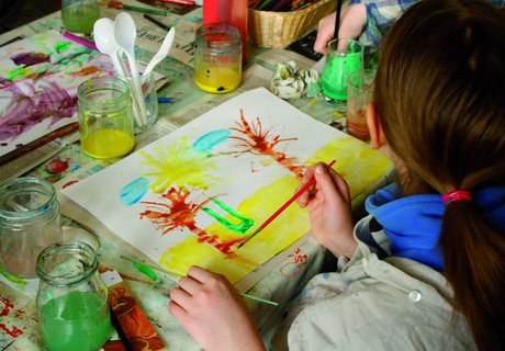 Ein Kind malt mit verschiedenen Farben und Pinseln ein Bild. Das Bild zeigt Palmen am Strand. Auf dem Tisch stehen Farbtöpfe und weitere Malutensilien.
