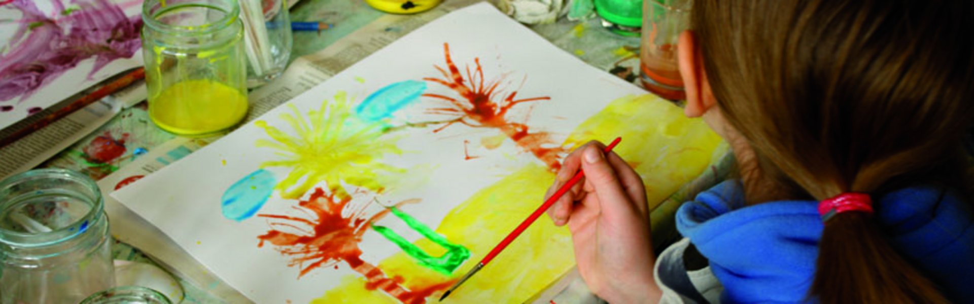 Ein Kind malt mit verschiedenen Farben und Pinseln ein Bild. Das Bild zeigt Palmen am Strand. Auf dem Tisch stehen Farbtöpfe und weitere Malutensilien.