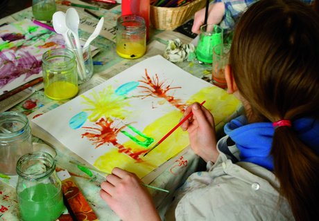 Ein Kind malt mit verschiedenen Farben und Pinseln ein Bild. Das Bild zeigt Palmen am Strand. Auf dem Tisch stehen Farbtöpfe und weitere Malutensilien.