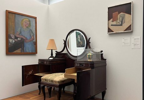 Auf der bunten Fotografie sieht man einen Ausstellungsraum. Vorne steht ein kleines Podest mit einer Rückwand und einer Seitenwand. An der Seitenwand hängt ein Gemälde. Es zeigt ein Mädchen mit blondem Haar. Sie trägt ein blaues Kleid mit einem grün-blauen Hemd darunter. Sie lehnt mit ihrem rechten Arm auf einem Tisch, auf dem verschiedene Schneckenhäuser liegen. Auf der Rückwand, oben rechts, hängt ein weiteres Gemälde mit verschiedenen Gegenständen. Auf dem Podest steht ein alter Schminktisch aus dunklem Holz. In der Mitte ist ein Spiegel. Vor dem Spiegel steht ein kleiner Hocker aus dem gleichen Holz mit einem grün verzierten Polster. Ganz hinten sieht man eine Wand. Dort hängt ein kleines Portrait von einem Mann, der eine Pfeife in der Hand hält. Er trägt eine Schiebermütze und einen Anzug mit Krawatte.