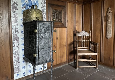 Auf dem Foto sieht man eine Bauernstube. In der Mitte steht ein Ofen, der Bilegger heißt. Der Ofen steht vor einer Wand mit blauen und braunen Fliesen. Neben dem Ofen steht ein Schaukelstuhl. Die Wände sind mit dunklem Holz bedeckt. So sieht eine typische Bauernstube aus.