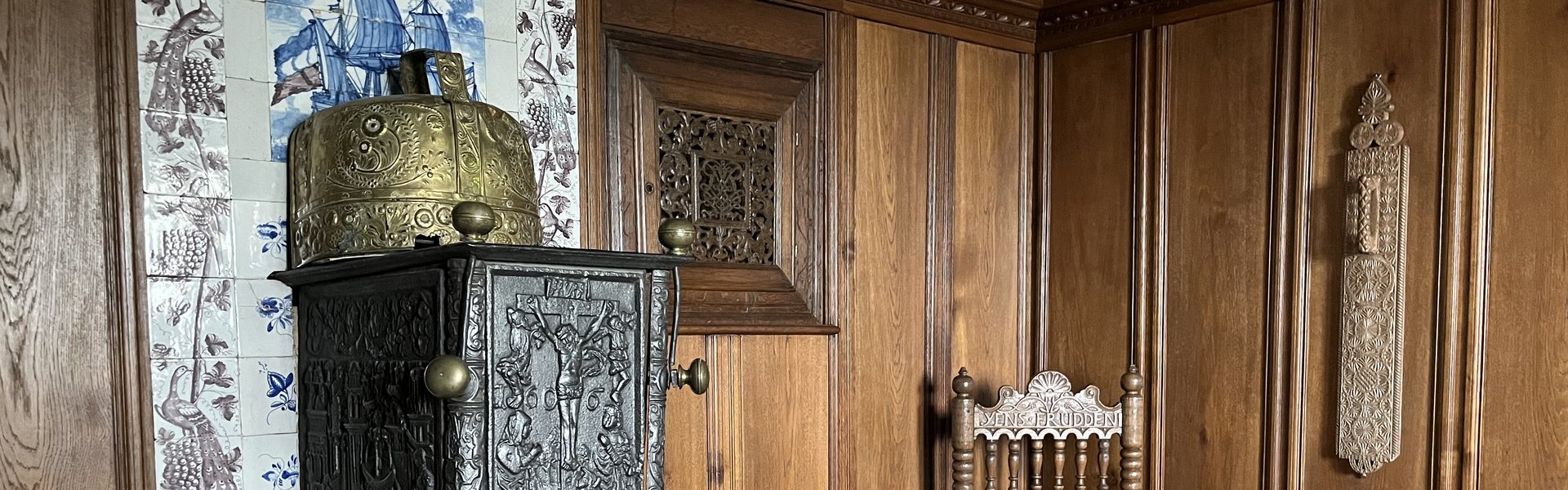 Auf der Fotografie sieht man die Einrichtung einer Bauernstube. Vor einer Wand mit blauen und braunen Fliesen steht ein Bilegger, ein Ofen. Rechts daneben befindet sich ein Schaukelstuhl. Die Wände sind mit dunklem Holz vertäfelt.