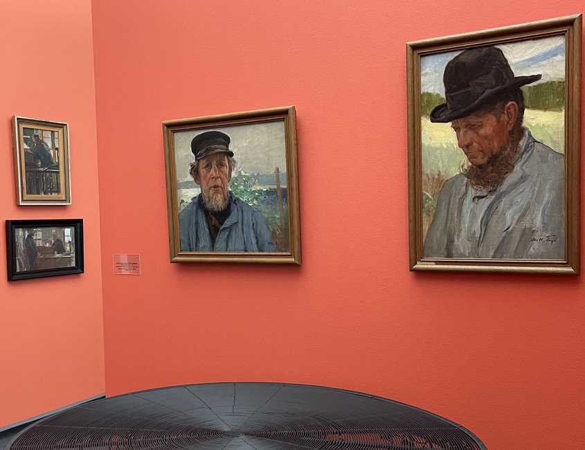 Auf dem Foto sind rechts zwei Gemälde mit Portraits eines Mannes zu sehen. Der Mann trägt eine dunkle Mütze und ein stabiles blaues Hemd. Er hat einen Bart. An der linken Wand hängen zwei weitere Bilder.