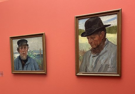 Auf dem Foto sehen Sie rechts zwei Gemälde. Beide zeigen ein Porträt von einem Mann. Ein Porträt ist ein Bild von einer Person. Der Mann trägt eine dunkle Mütze und ein stabiles blaues Hemd. Er hat einen Bart. An der linken Wand hängen zwei weitere Bilder.