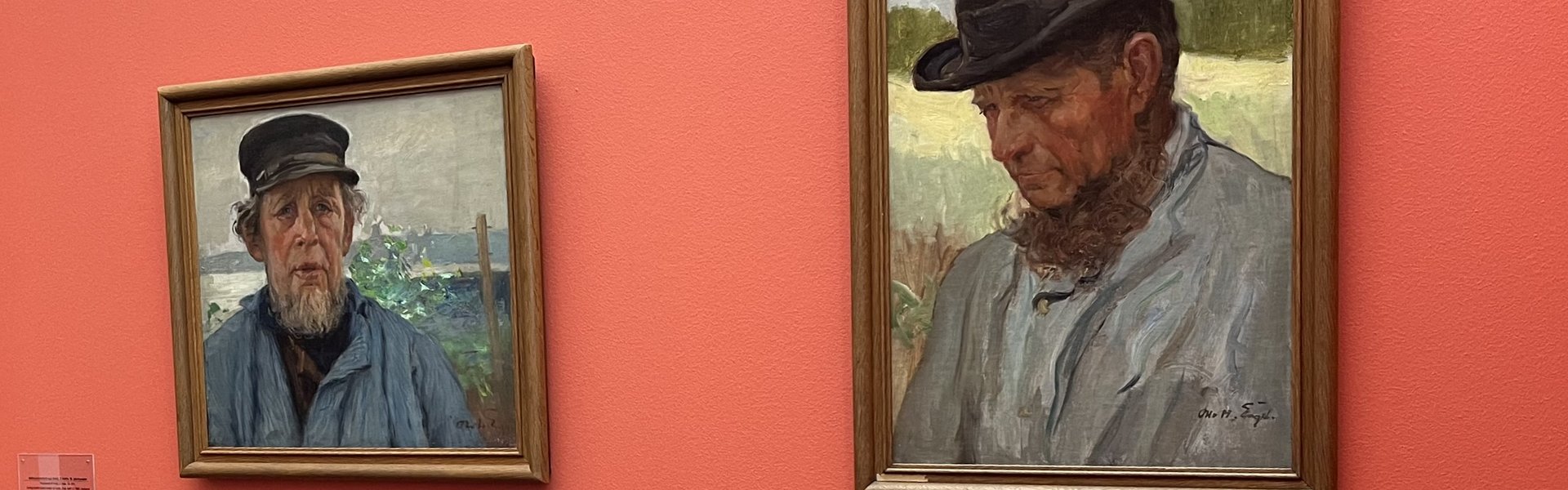 Auf dem Foto sind rechts zwei Gemälde mit Portraits eines Mannes zu sehen. Der Mann trägt eine dunkle Mütze und ein stabiles blaues Hemd. Er hat einen Bart. An der linken Wand hängen zwei weitere Bilder.