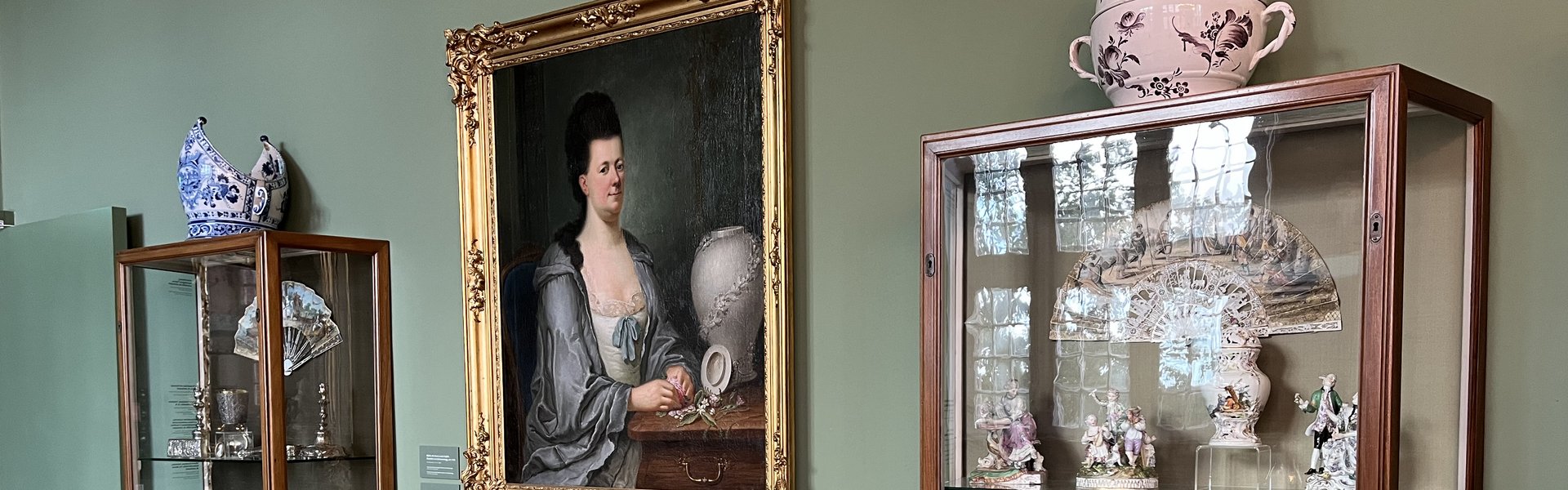 Die Fotografie zeigt in der Mitte ein Gemälde. Auf dem Gemälde sieht man eine Frau. Es ist ein Brustbild von einer Adligen, die einen Morgenmantel trägt. Ein Brustbild ist ein Bild, auf dem nur der obere Teil einer Person zu sehen ist. Vor ihr steht eine Vase. Unter dem Gemälde steht ein kleiner Tisch. Auf dem Tisch steht ebenfalls eine Vase. Rechts und links vom Tisch stehen Glasvitrinen. In diesen Vitrinen befinden sich verschiedene Objekte.