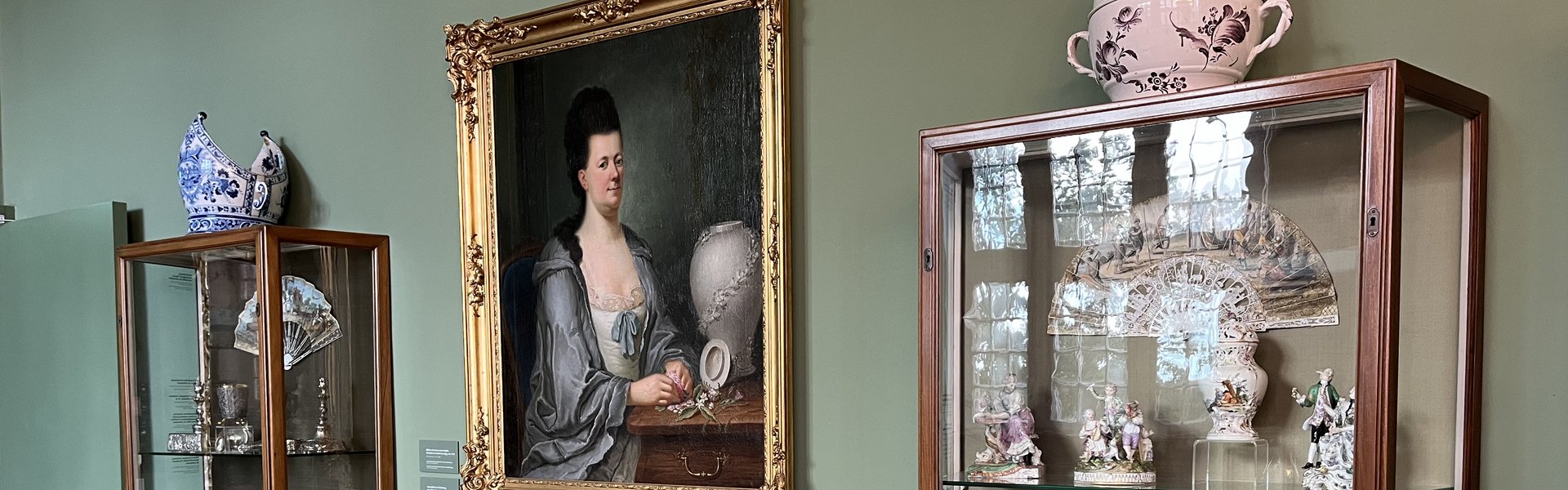 Die Fotografie zeigt in der Mitte ein Gemälde, auf dem eine Frau zu sehen ist. Das Brustbild zeigt eine Adelige im Morgenmantel. Vor ihr steht eine Vase. Unter dem Gemälde steht ein kleiner Tisch. Auf dem Tisch steht auch eine Vase. Rechts und links daneben stehen Glasvitrinen. In ihnen befinden sich verschiedene Objekte.  