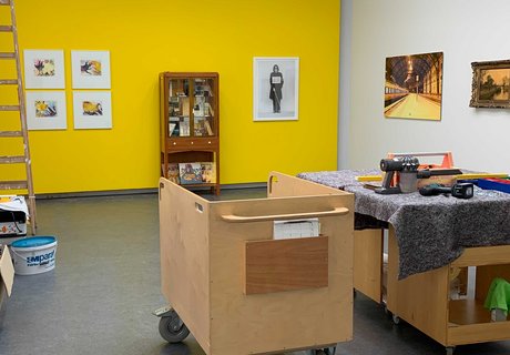 Auf dem Foto ist ein gelber Ausstellungsraum zu sehen. An der gelben Wand hängen unterschiedliche Bilder und in der Mitte steht ein Vitrinenschrank. Im Vordergrunde steht ein kleiner Transportwagen und daneben ein mobiler Arbeitstisch mit unterschiedlichen Werkzeugen. Links im Bild ist eine Holzleiter und verschiedene Malermaterialien. 