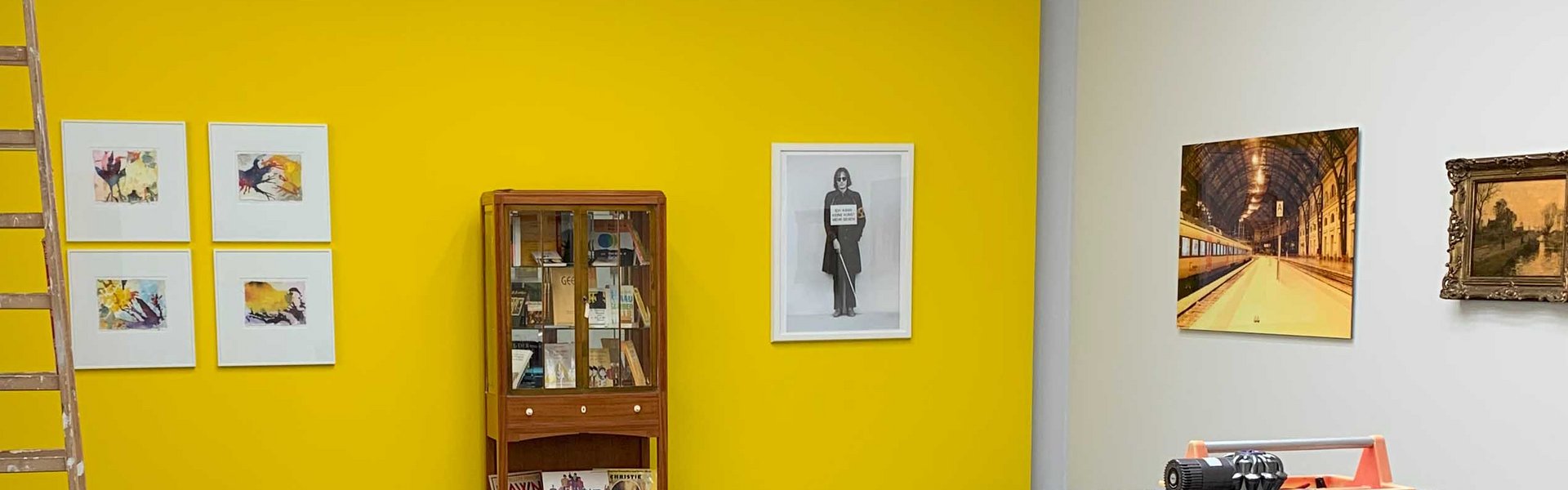Auf dem Foto ist ein gelber Ausstellungsraum zu sehen. An der gelben Wand hängen unterschiedliche Bilder und in der Mitte steht ein Vitrinenschrank. Im Vordergrunde steht ein kleiner Transportwagen und daneben ein mobiler Arbeitstisch mit unterschiedlichen Werkzeugen. Links im Bild ist eine Holzleiter und verschiedene Malermaterialien. 