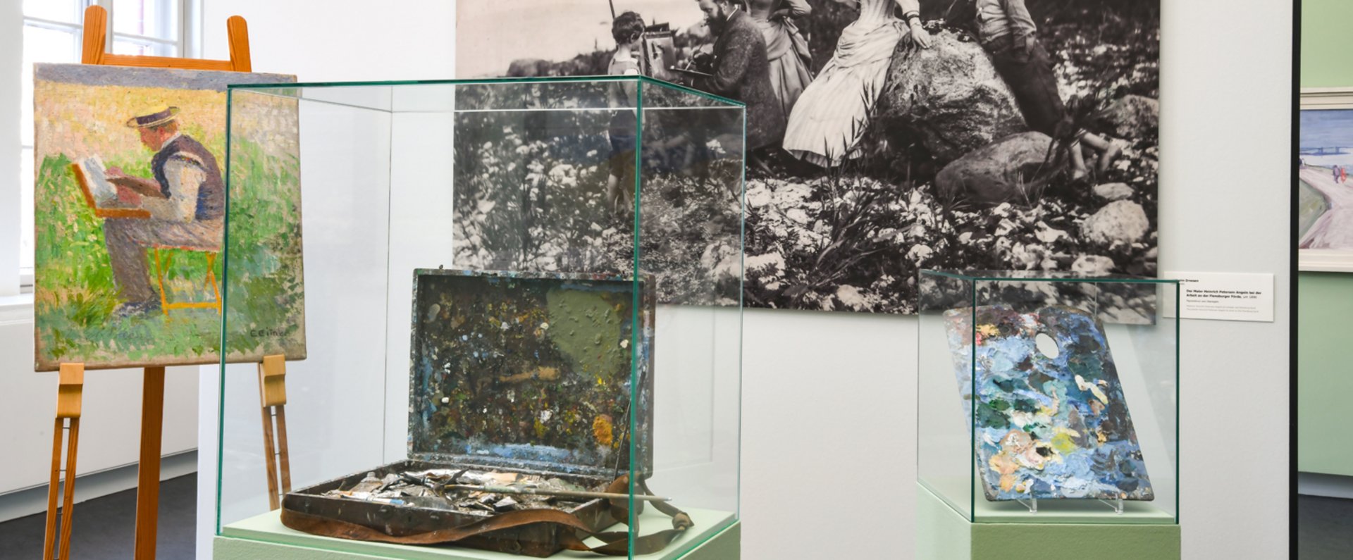 Ein großes schwarz-weiß Foto hängt an der Wand. Davor steht ein Gemälde auf einem Ständer. Daneben sind alte Pinsel und Farbe in einem Glaskasten.