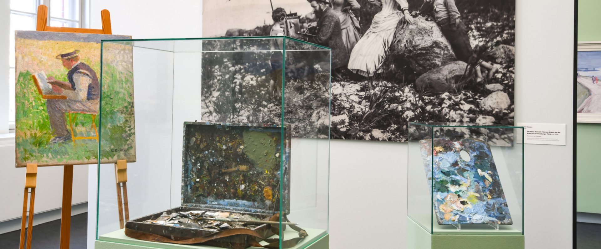 Ein großes schwarz-weiß Foto hängt an der Wand. Davor steht ein Gemälde auf einem Ständer. Daneben sind alte Pinsel und Farbe in einem Glaskasten.