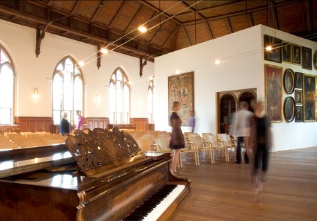 Det er auditoriet på Museumsberg Flensburg. Der kan man afholde koncerter. Der afholdes også andre arrangementer, f.eks. når der er en ny udstilling, der skal fejres. Der er mange stole og mennesker, der bevæger sig rundt. Foran ses et klaver. I baggrunden ses det parisiske værelse. Det er et rum, der er indrettet som et kunstværk.