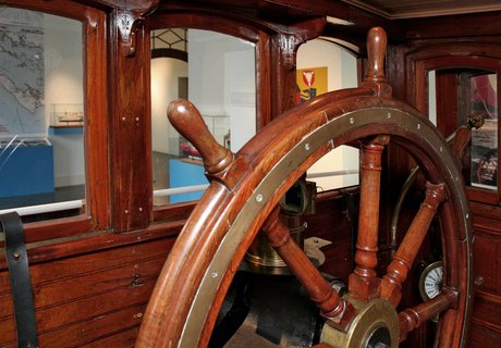 Das Foto zeigt den Blick aus dem Steuerhaus HABICHT in die Ausstellung FÖRDESCHIFFAHRT im Flensburger Schifffahrtsmuseum. Im Steuerhaus ist ein großes Ruder bzw. Steuerrad mit dem das Schiff früher gesteuert worden ist.